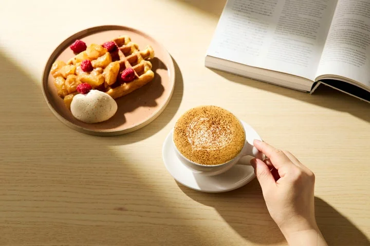 ワッフルプレートとコーヒーカップが置かれたテーブルで、本を読みながらコーヒーを楽しむ手元