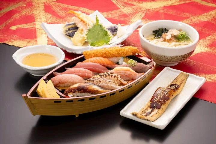 船型の器に盛られた握り寿司と天ぷら、うどん、穴子焼きが並ぶ食事風景