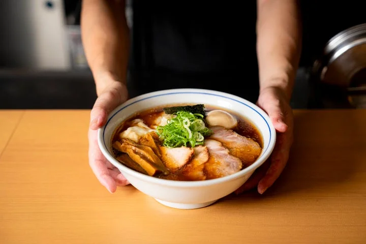 両手でラーメン丼を差し出す店員と、チャーシューやネギが乗った醤油ラーメン