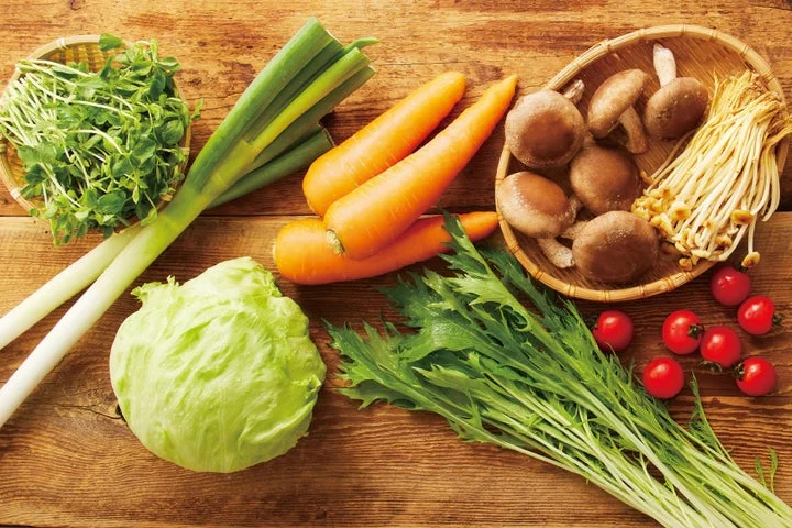木製の台の上に並べられた長ねぎ、にんじん、白菜、きのこ、豆苗、トマトなどの生野菜