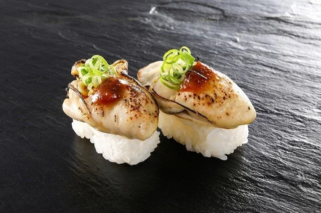 炙り蒸し牡蠣南蛮味噌。黒い皿に2貫の握り寿司が並び、牡蠣の上に南蛮味噌とネギが乗っている。