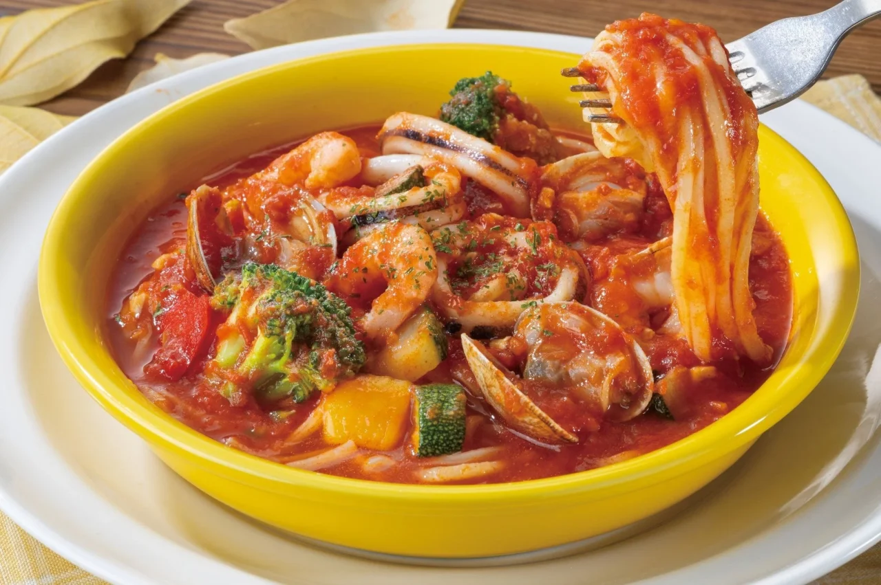 魚介たっぷりトマトスープパスタ。黄色い器にトマトスープと魚介、野菜が盛り付けられ、フォークでパスタが持ち上げられている