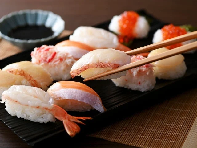黒い長方形の皿に盛られた生海老やいくらなど複数のネタのにぎり寿司と、それを掴む箸