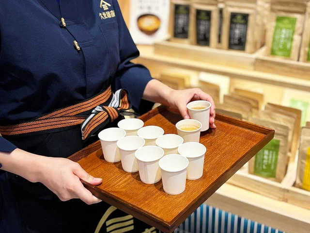 店舗スタッフが出し汁の試飲用紙コップを木製トレイに乗せて提供している様子