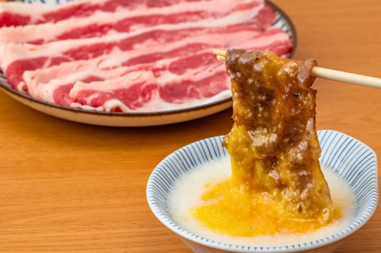 とろ玉すき焼きカルビ。卵黄ととろろのタレにくぐらせた焼きカルビを箸で持ち上げ、背景に生カルビが写る。