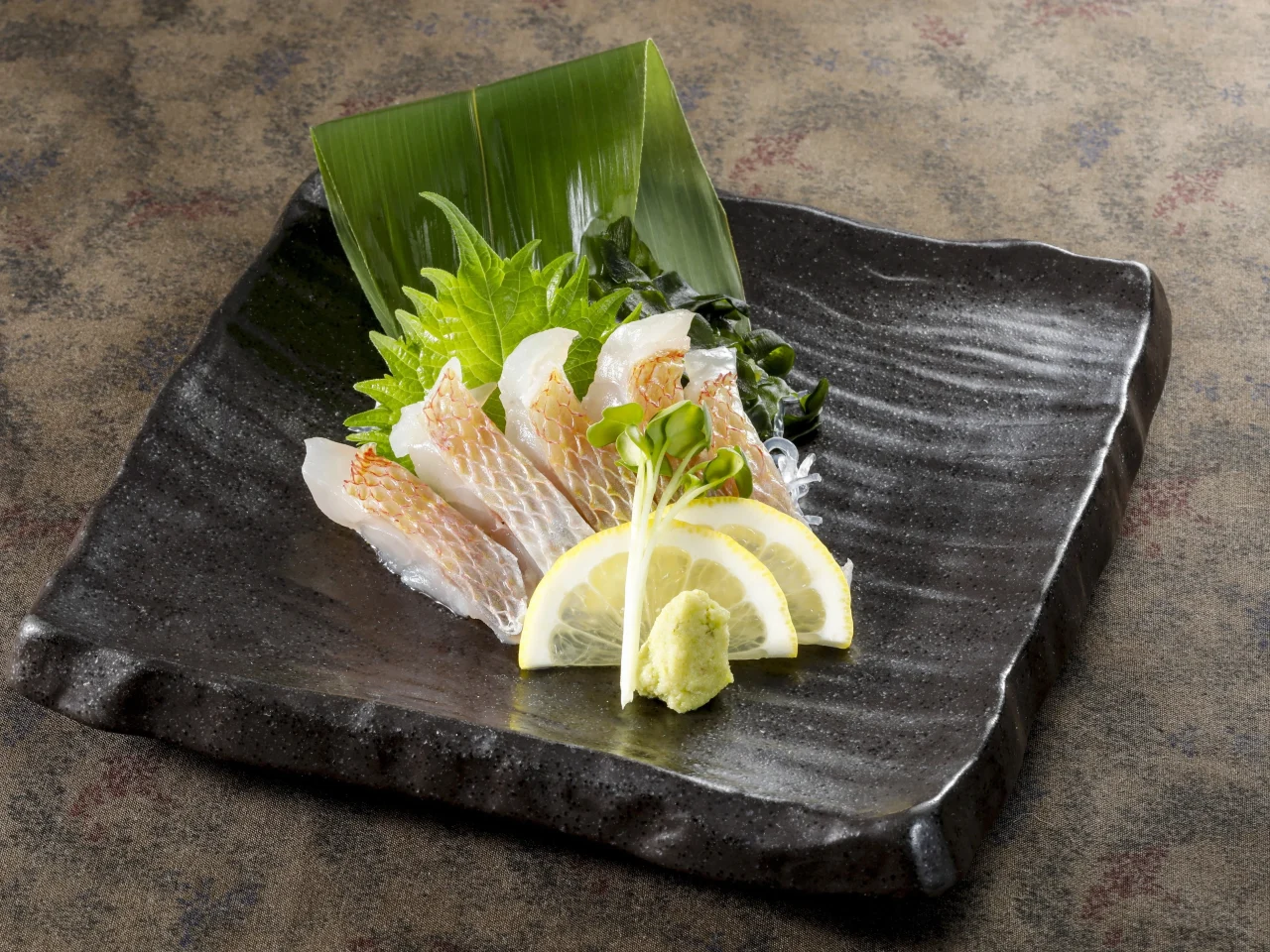 き鯛湯霜お造り。黒い長皿に薄切りの魚とレモン、薬味を添えて盛り付けた様子。
