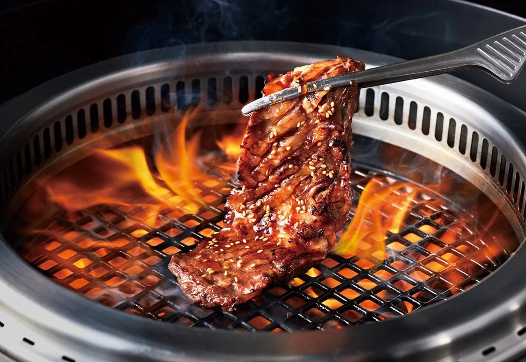 焼肉きんぐの炭火焼肉。網の上で焼かれた肉をハサミで持ち上げ、炎と煙が立ち上る様子。