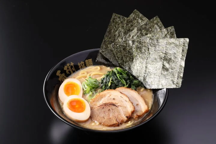 黒い丼に盛られた家系ラーメン。半熟卵、チャーシュー、ネギ、青菜、海苔が乗っている
