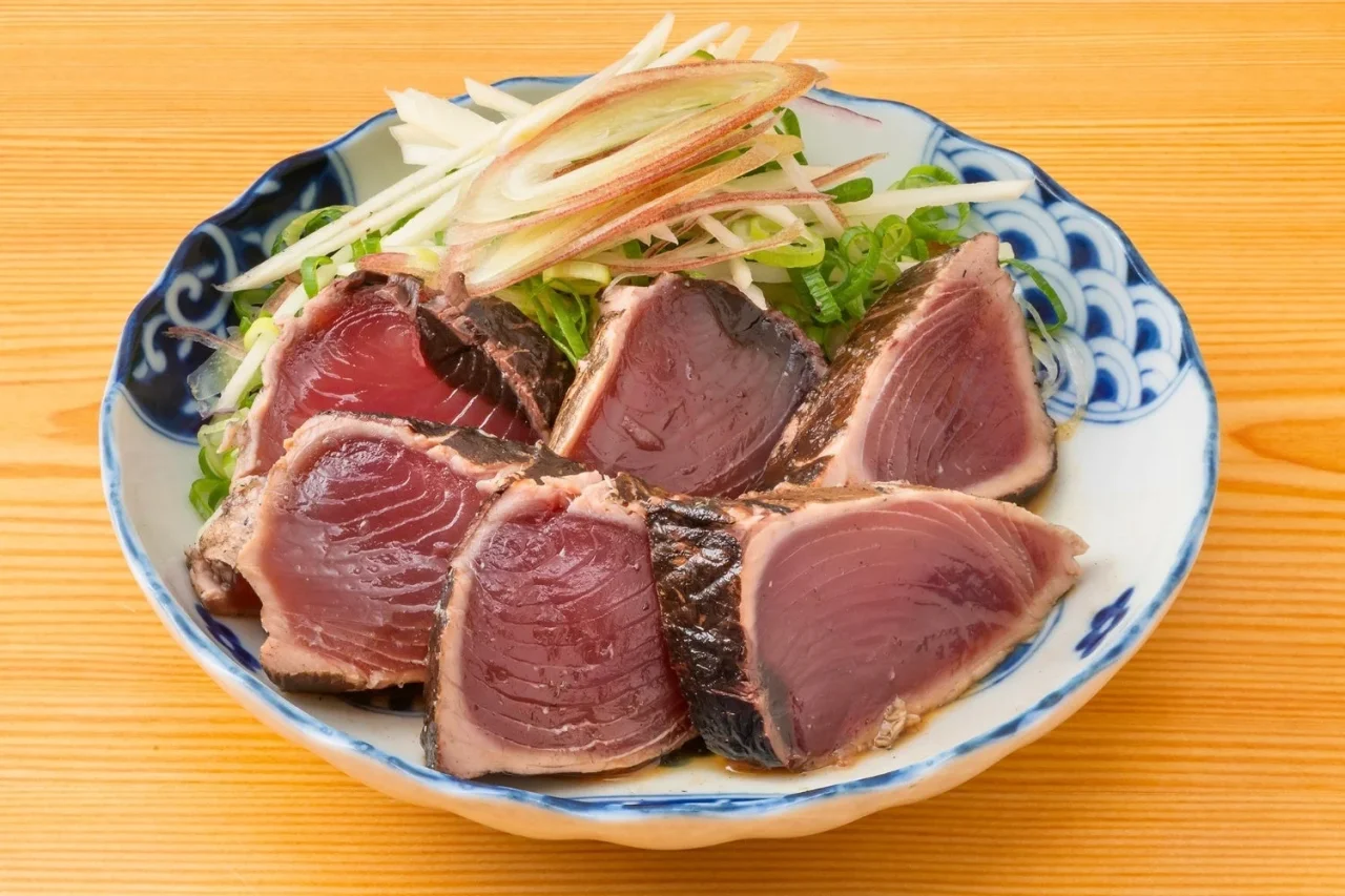 わら焼き鰹たたき。焼き色がついた鰹のたたきを、薬味とともに青白の皿に盛り付けた様子。