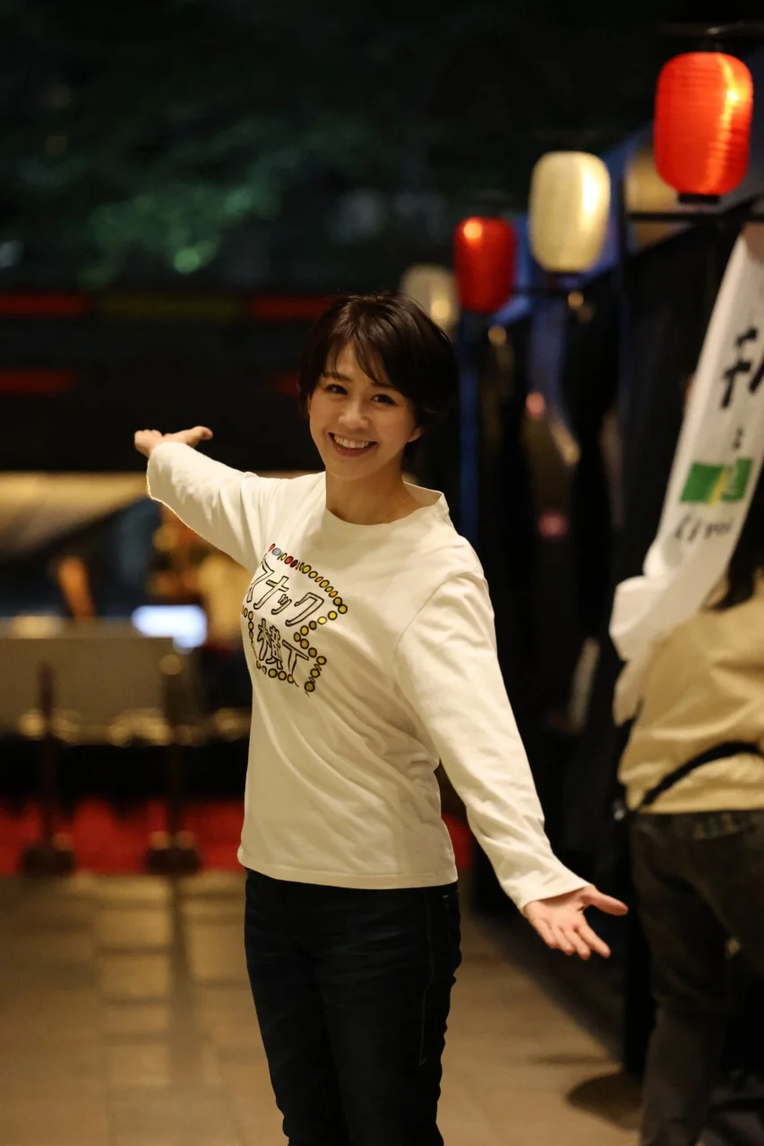 スナック横丁のロゴ入りTシャツを着た五十嵐真由子代表が、夜の屋外で両手を広げて笑顔で立っている