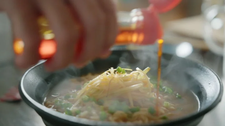 黒い丼に盛られたラーメンに、白髪ネギとネギをトッピングし、ラー油を注ぐ様子