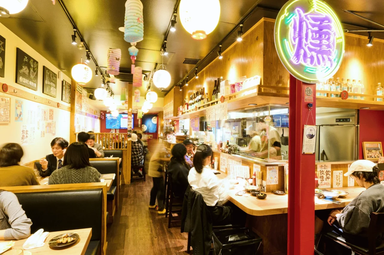 とりとんくん・燻し家もっくんの店内。客席とカウンターに客が座り、提灯やネオンが飾られた活気ある空間。