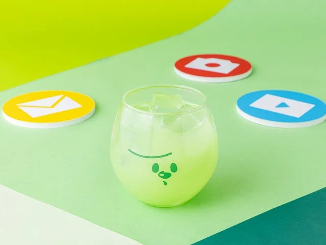 メメルの顔が描かれたグラスに入った緑色の炭酸ドリンクと、アプリ風アイコンのコースター