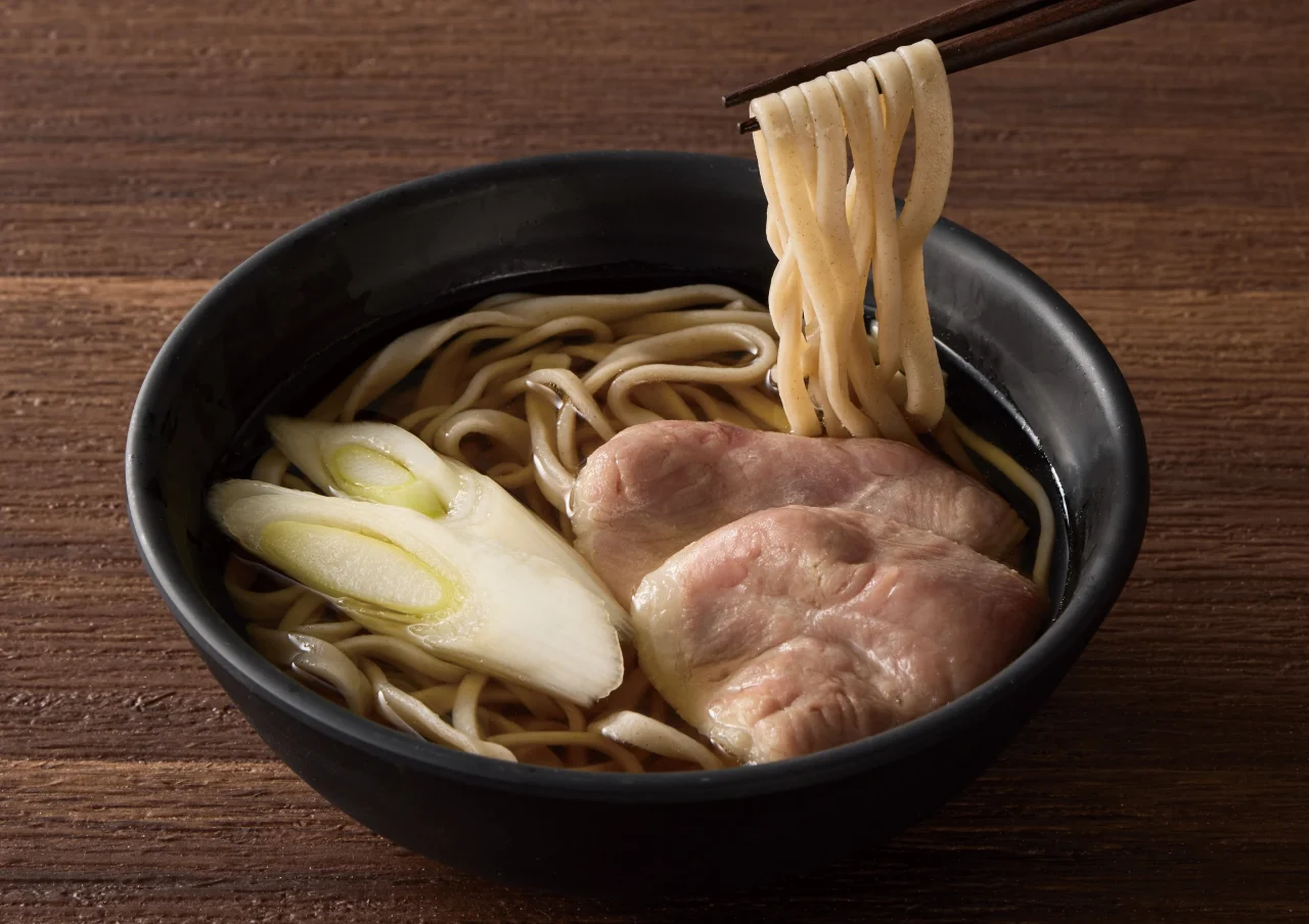 鴨そば。黒い丼にそば、鴨肉、葱が盛り付けられ、箸でそばが持ち上げられている
