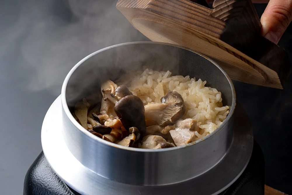 鶏と「ひらたけ」の秋香る釜めし。蓋を開けた釜から湯気が立ち、ひらたけと鶏肉が乗ったご飯が見える。