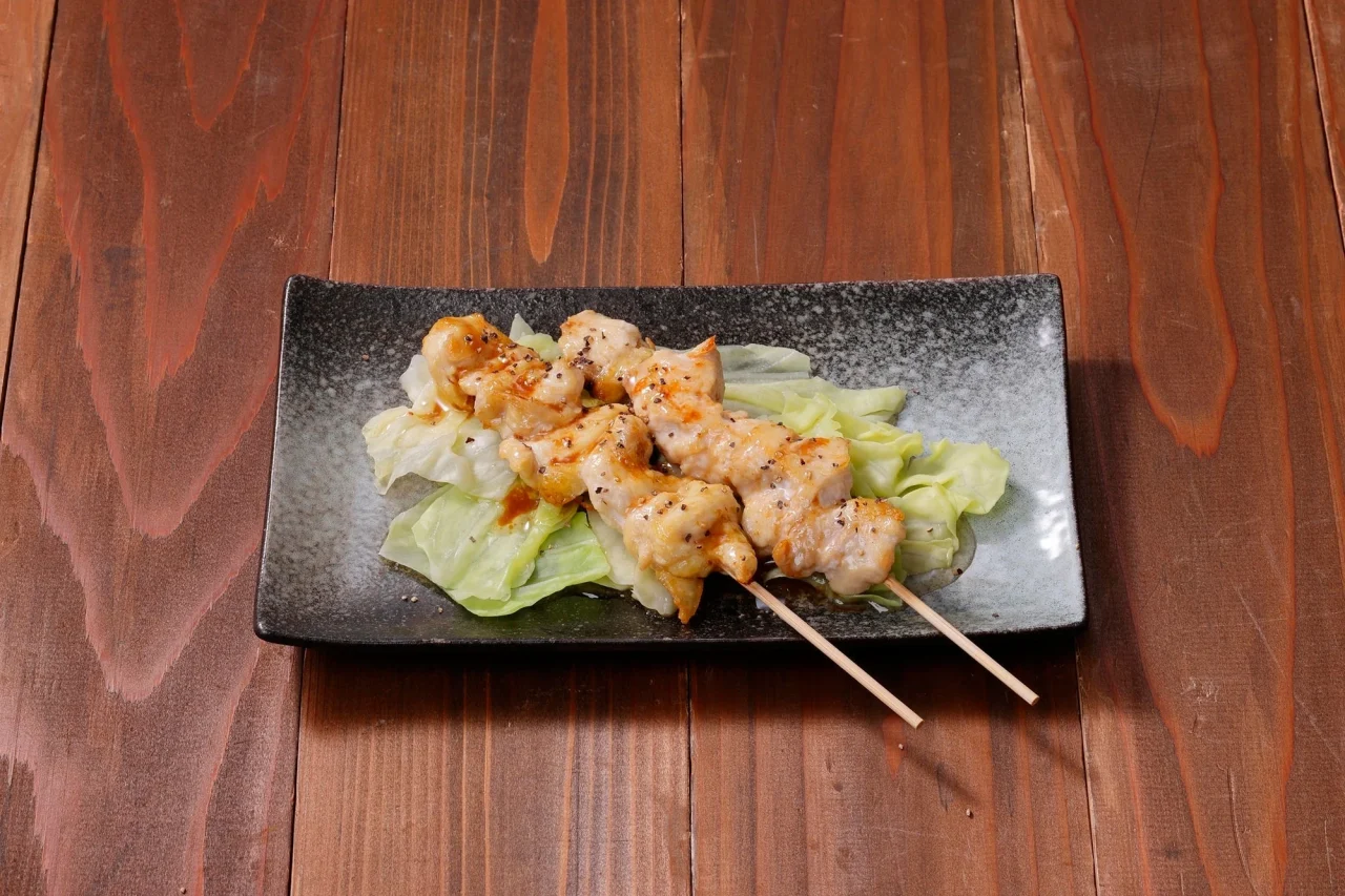 キャベツを敷いた黒い長皿に、ソースをかけた焼き鳥の串が2本置かれた肩肉のオイルソース焼