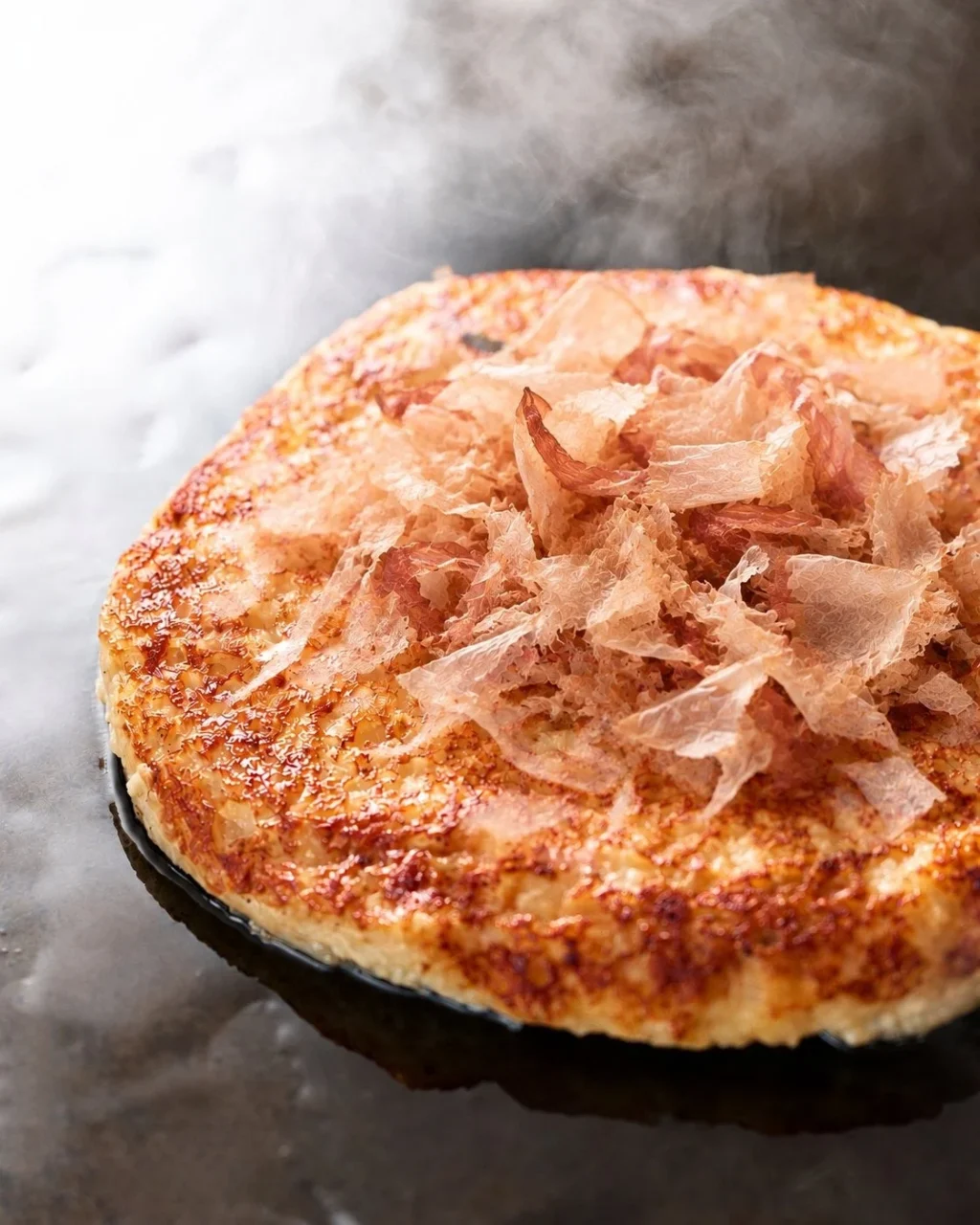 鉄板の上で焼かれた円形のもんじゃ焼き。表面は焦げ目がつき、中央に削り節が散らされている。