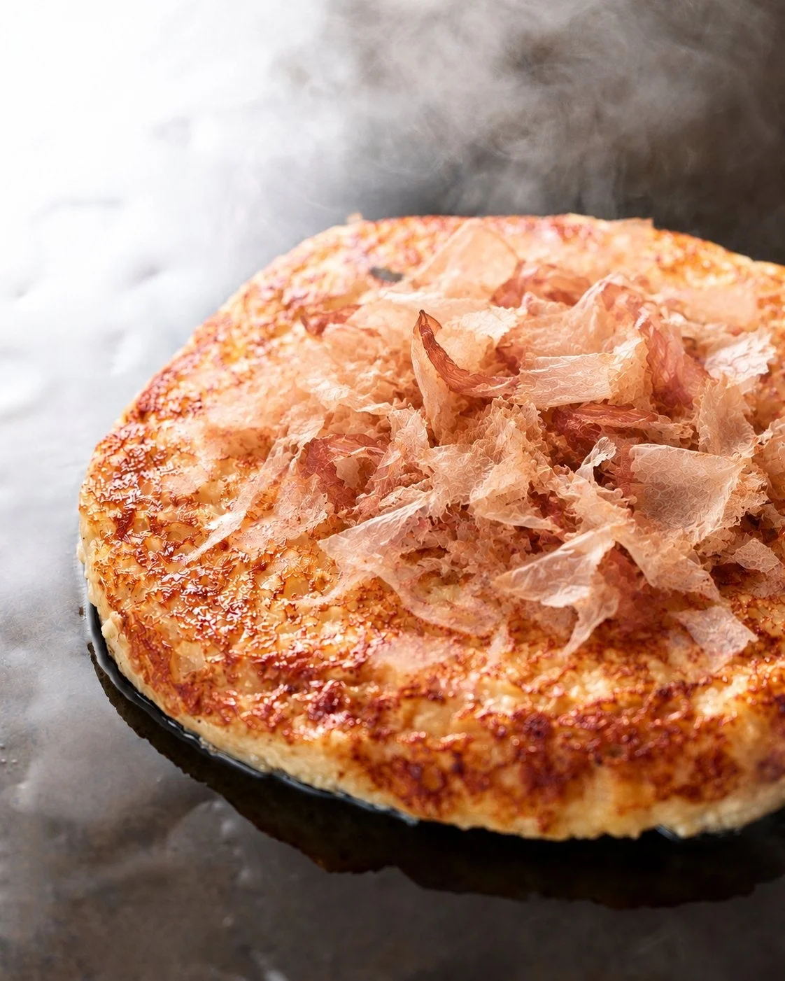 鉄板の上で焼かれた円形のもんじゃ焼き。表面は焦げ目がつき、中央に削り節が散らされている。