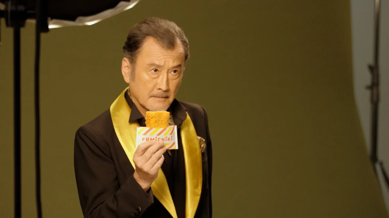 吉田鋼太郎が「FAMITCHIKI」の文字が書かれたカードに挟まれたファミチキを手にするTVCMの一場面
