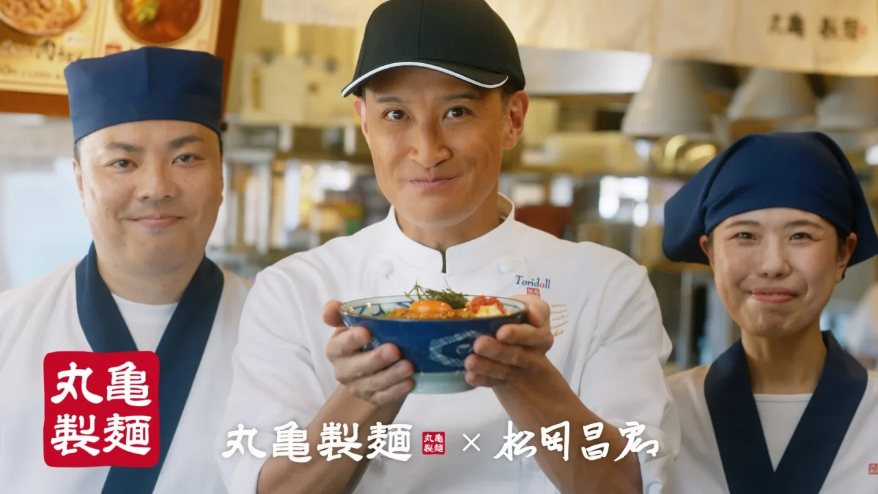 丸亀製麺「肉辛まぜうどん」TVCMカット。松岡昌宏さんがうどん丼を手に持ち、麺職人と並んで笑顔で写っている。