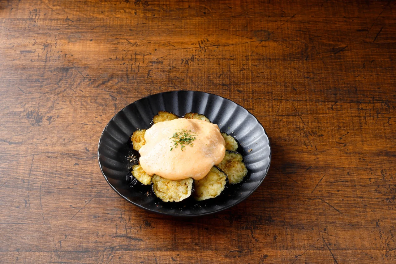 揚げナスの明太チーズまみれ。黒い皿に揚げナスとチーズソースを盛り付けた料理。