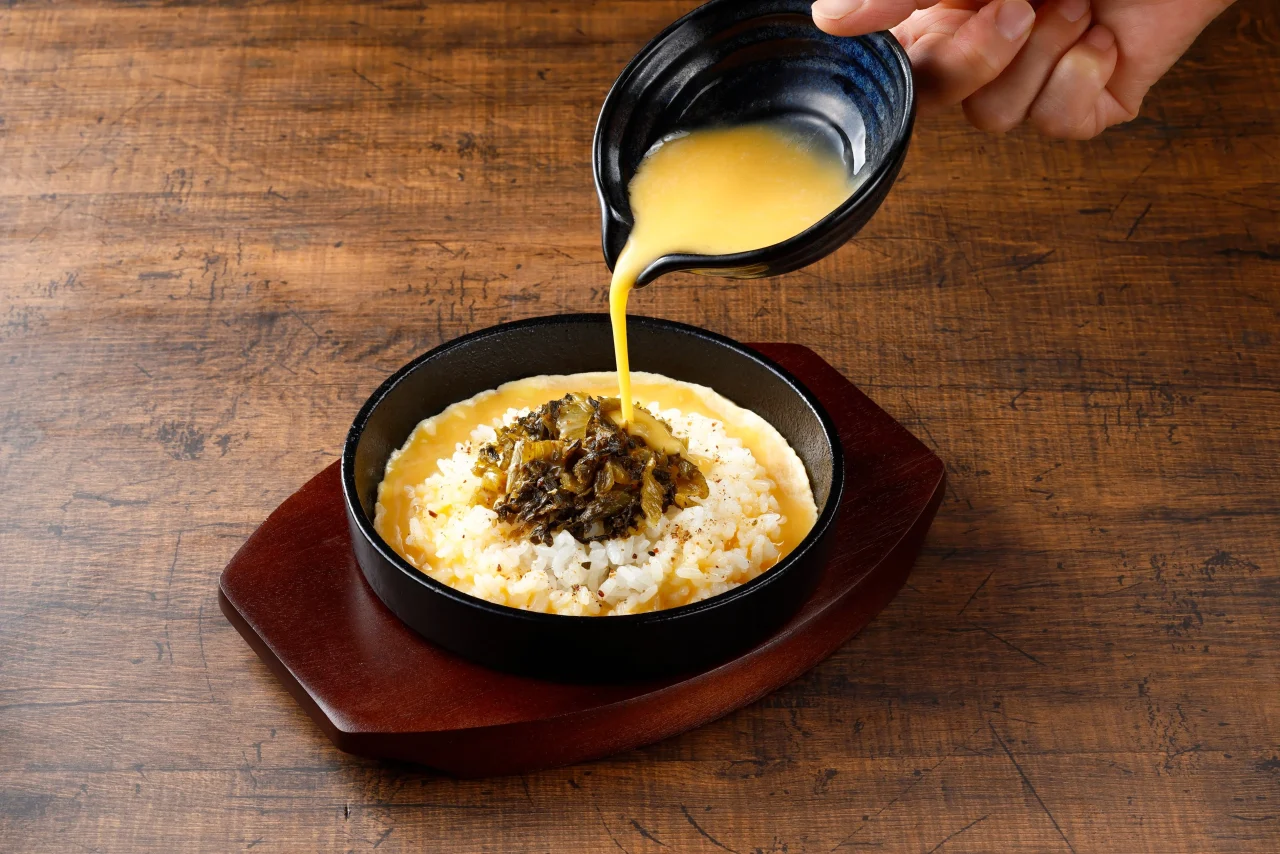 鉄板 高菜焼きめし。黒い器に盛り付け、高菜を載せたご飯に黄色いソースを注ぐ様子。