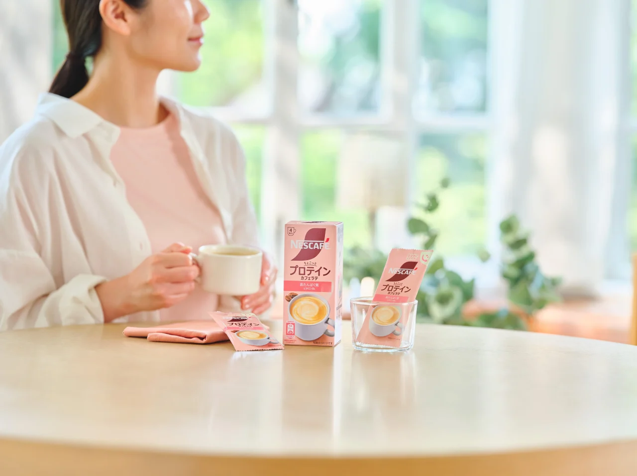 女性がカップを持ち、卓上にネスカフェ プロテイン カフェラテの箱とスティックが並ぶ
