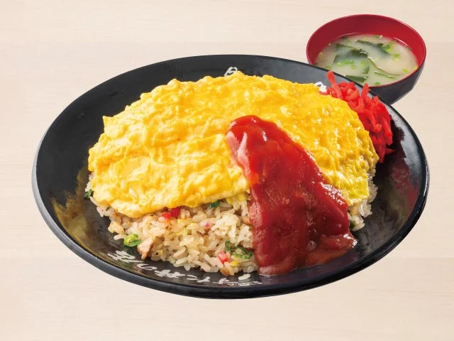 黒い皿に盛られたオムチャーハン。ふわとろのオムレツとケチャップ、スープが添えられている。
