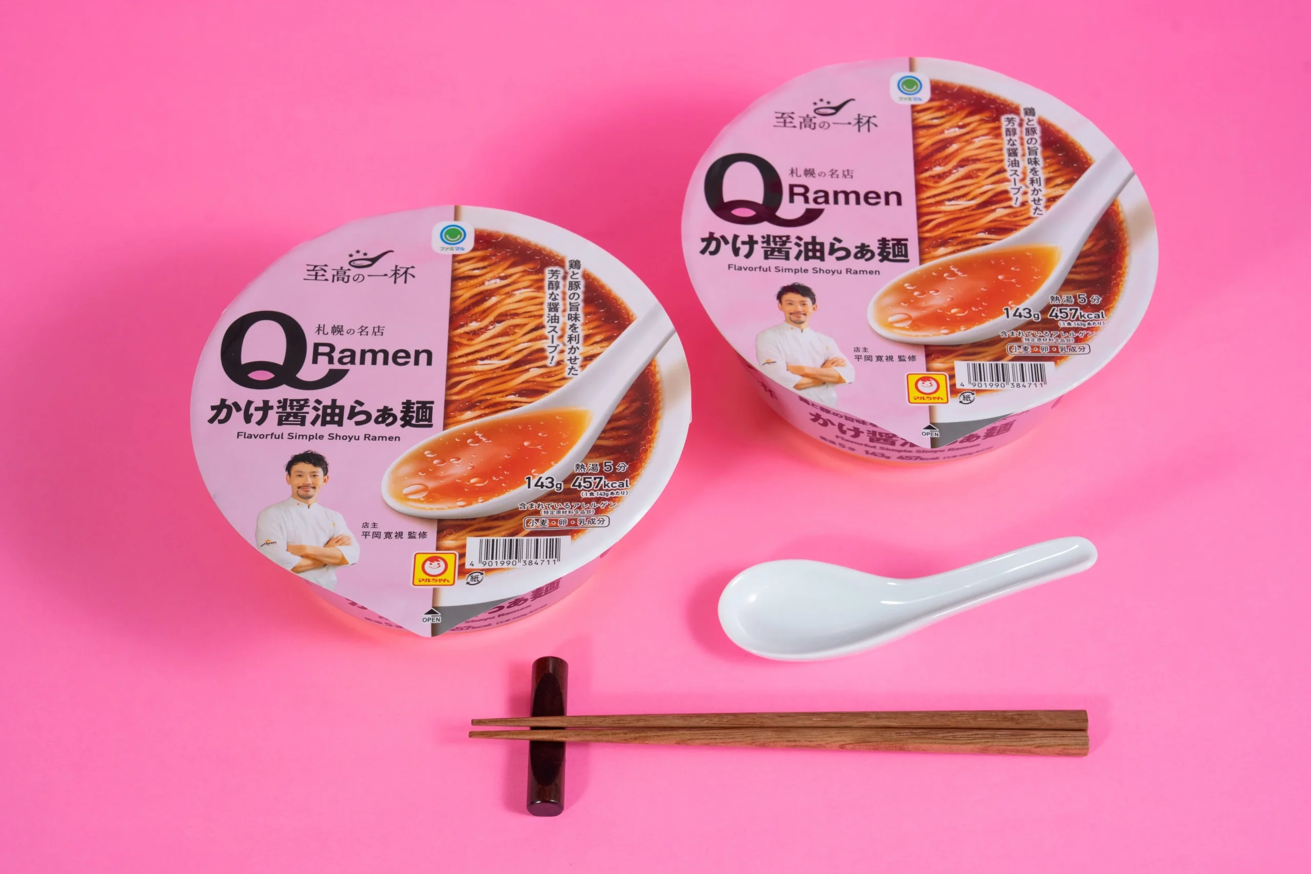 ピンクの背景にQRamen かけ醤油らぁ麺のカップ麺2個とスプーン、箸が置かれた様子