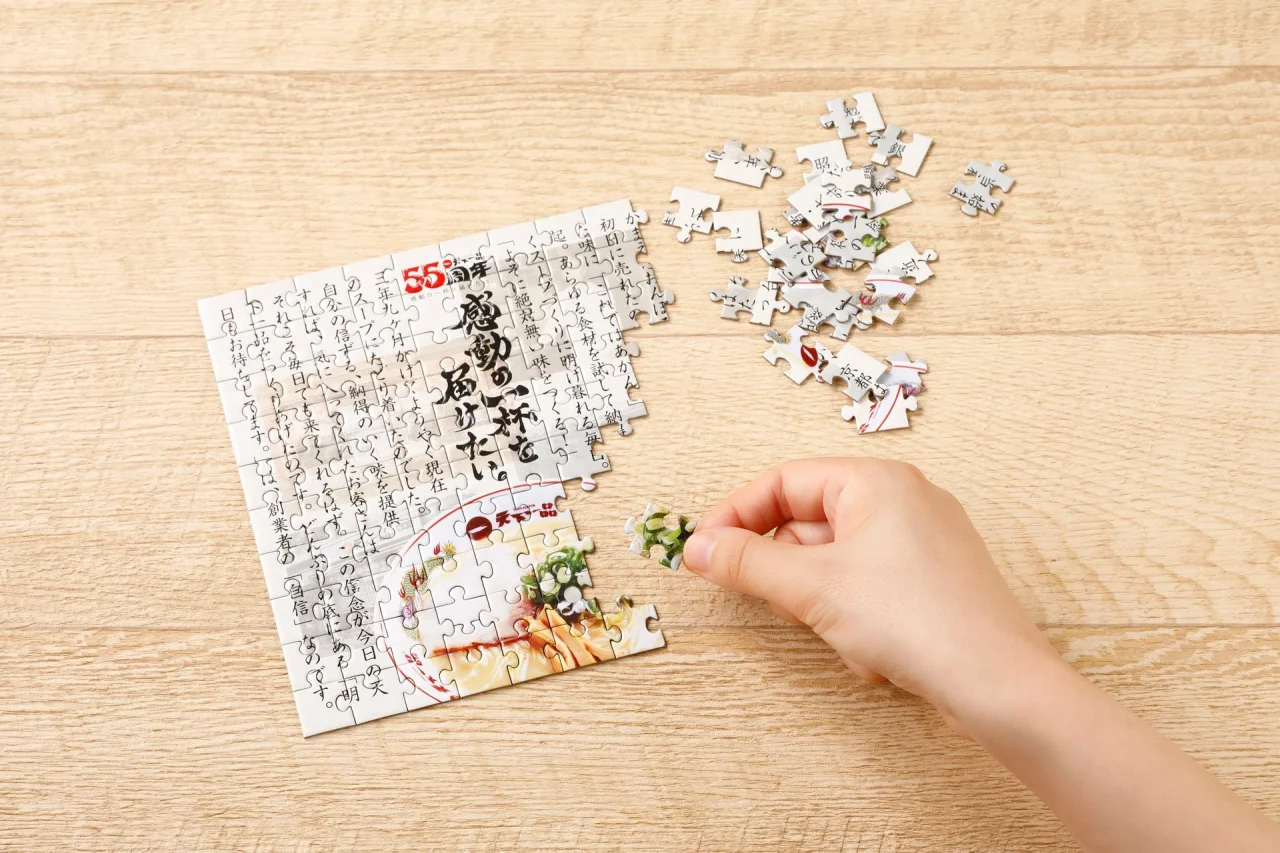 【B賞】ジグソーパズル。木目の机で子どもの手がピースをはめている