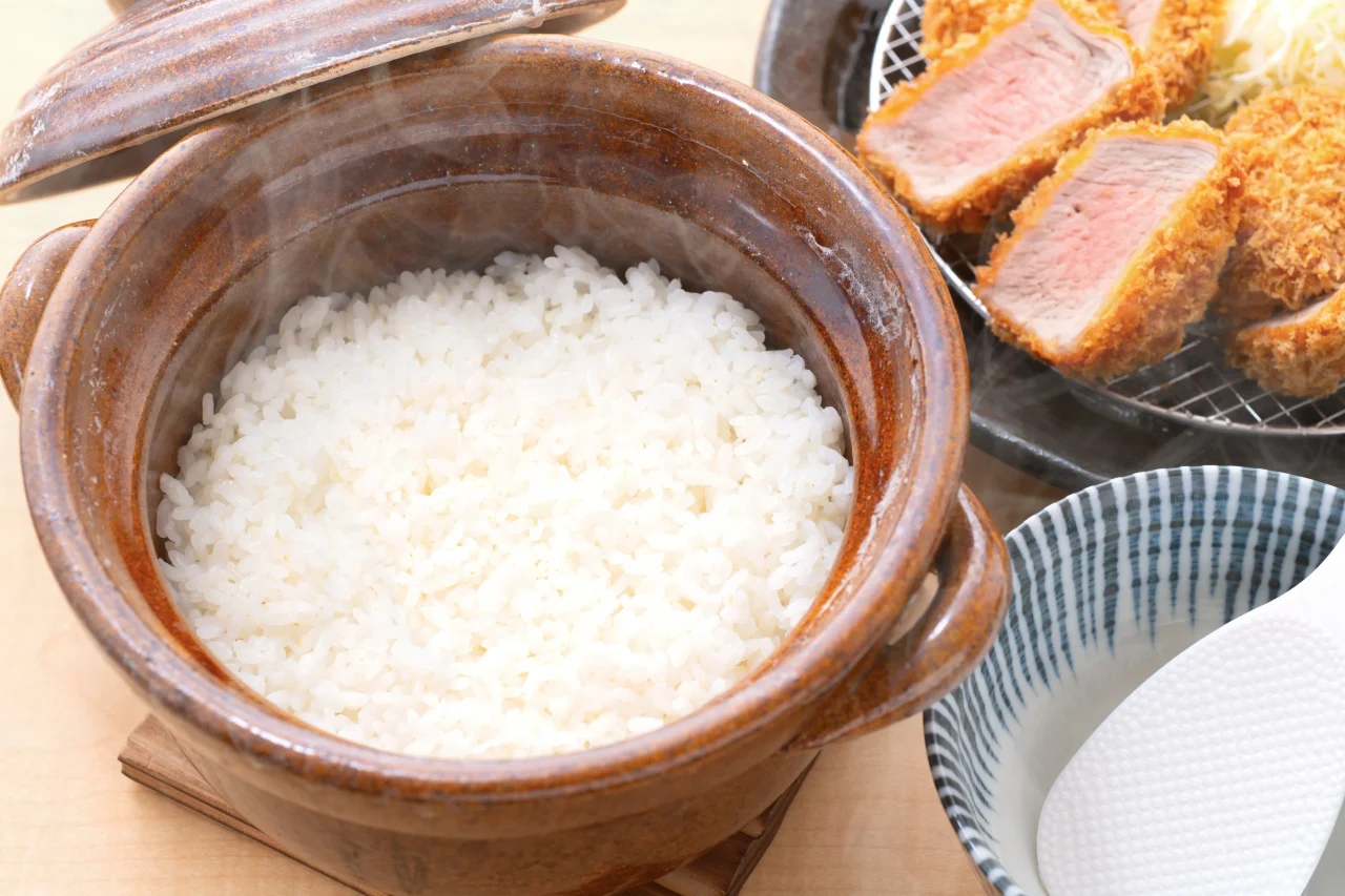 蓋を開けた土鍋ごはん。白米から湯気が立ち、奥にとんかつが見える
