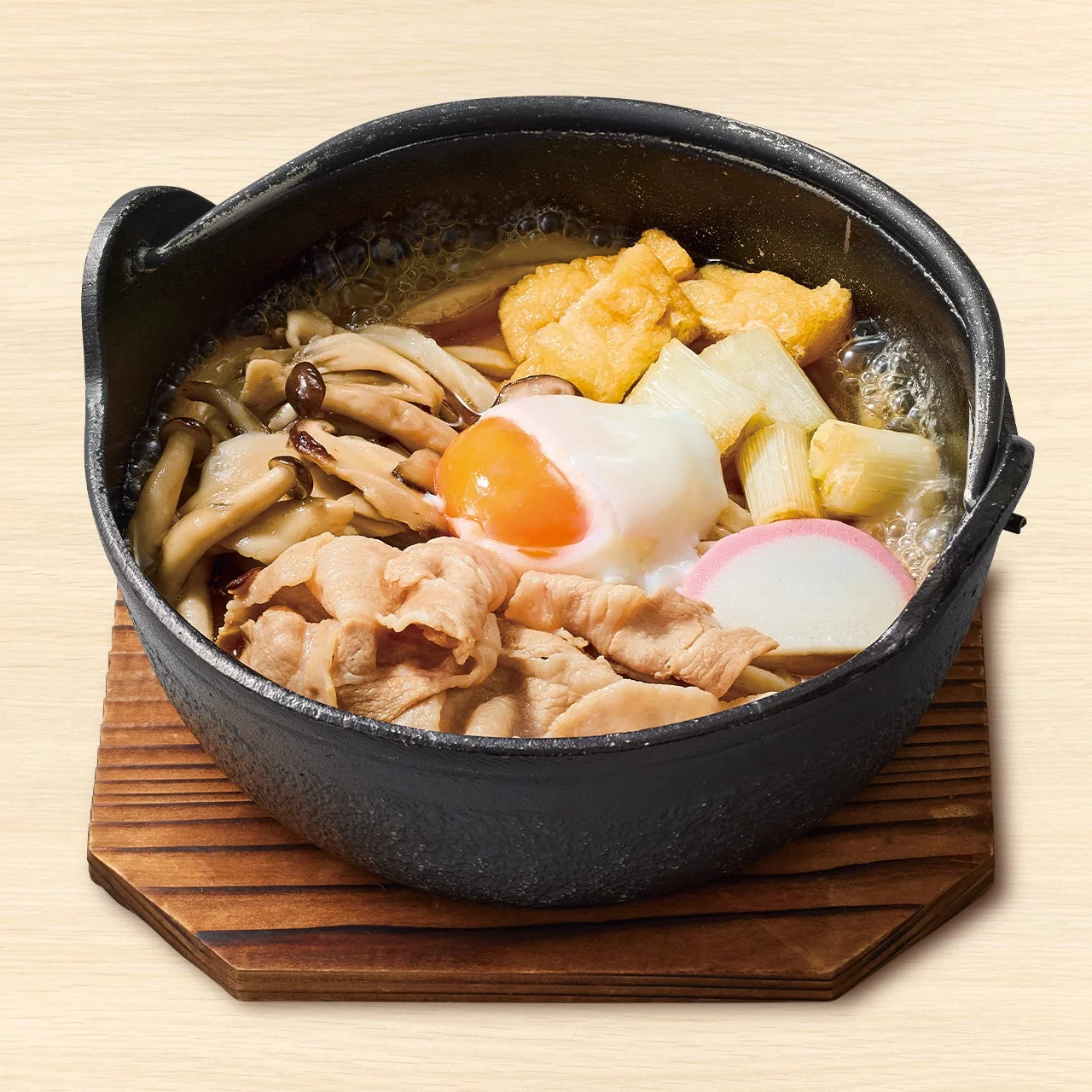 木の鍋敷きにのせた黒い鍋の肉鍋焼きうどん。豚肉、きのこ、温玉入り