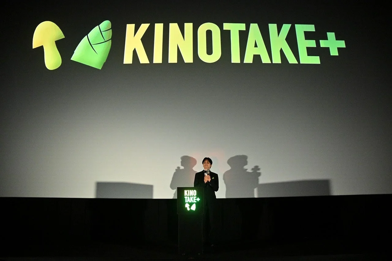 KINOTAKE+のロゴが映るスクリーン前に立つタキシード姿の男性