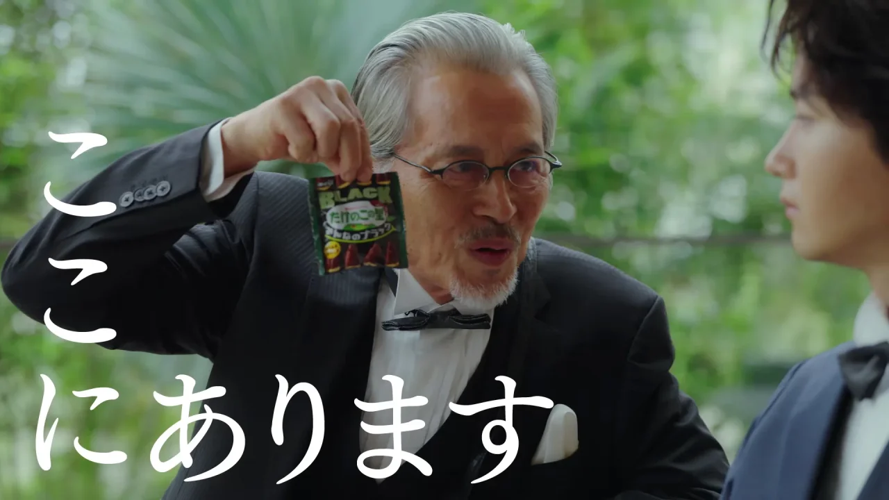 黒いパッケージの菓子袋を掲げる、眼鏡にタキシード姿の年配男性とCM場面