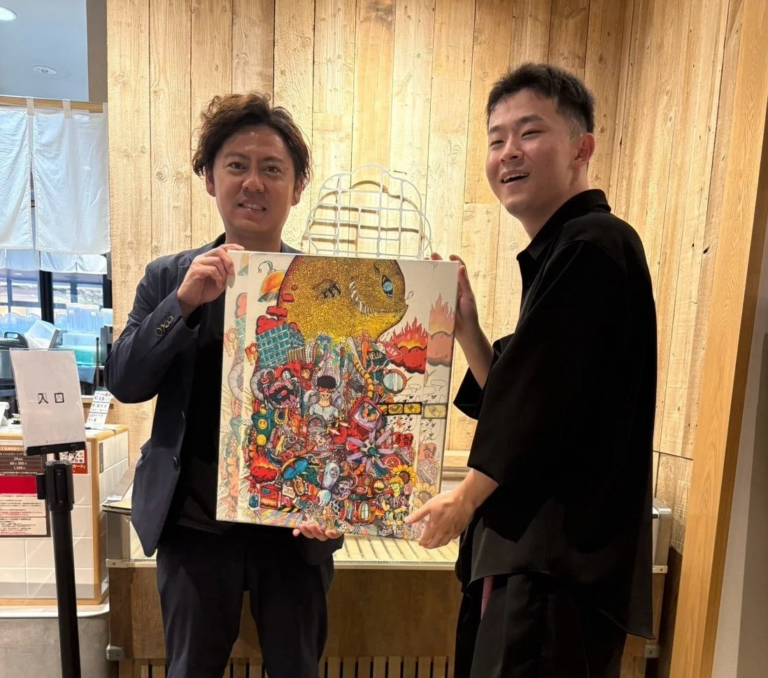 安部真良社長(左)とMUSASHI(右)がカラフルな絵画を持つ