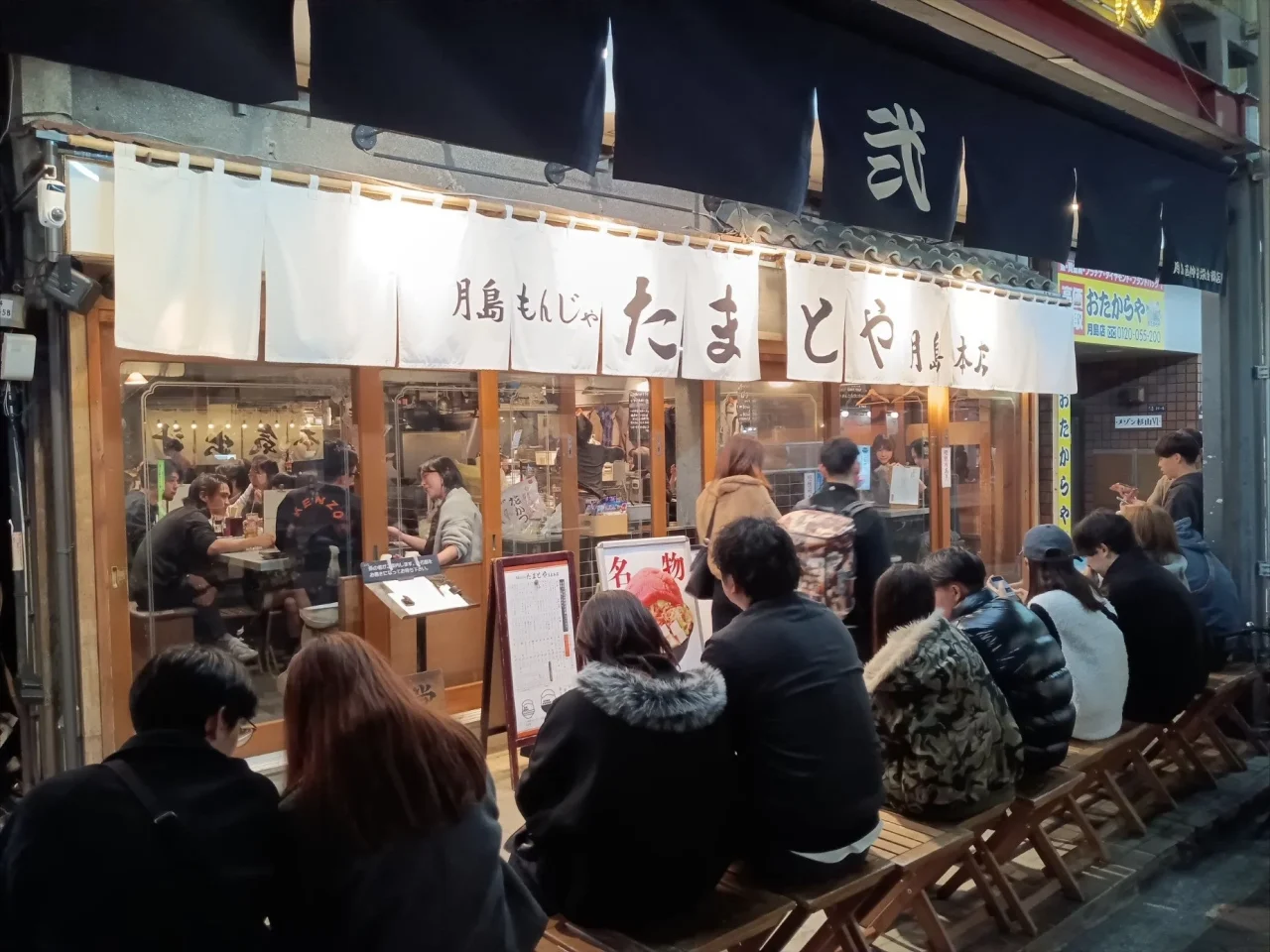 夜の月島もんじゃ たまとやの店頭。暖簾と、外のベンチに並ぶ客