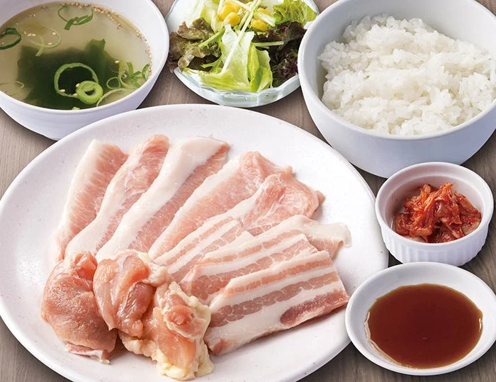 お手頃焼肉定食。生の豚肉・鶏肉の皿にご飯、スープ、サラダ、キムチ、タレ