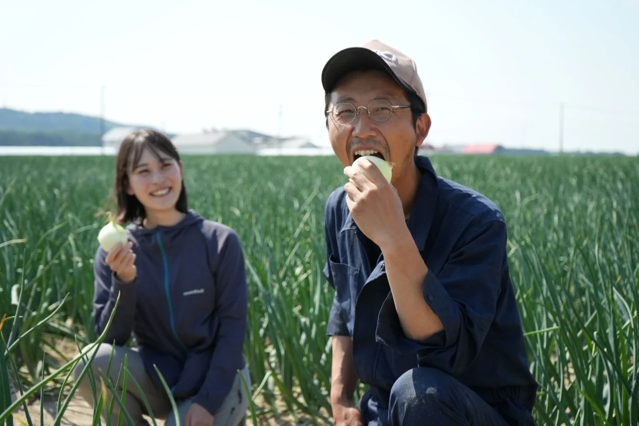畑でタマネギを丸かじりする男性と、タマネギを持って笑う女性