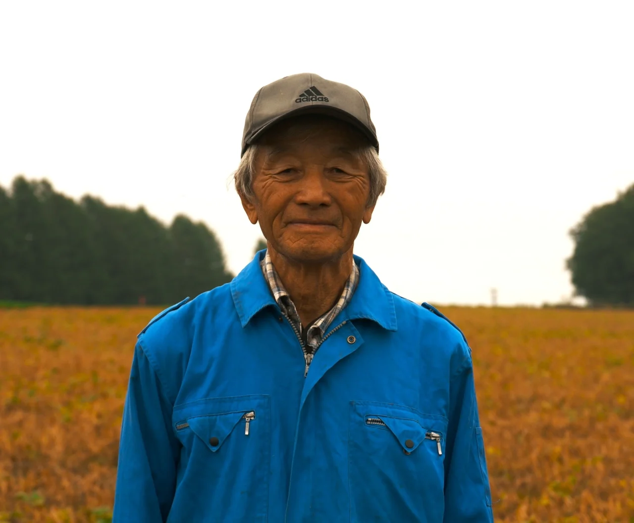 勇退する生産者・辻野さんが畑の前で微笑む、青い作業着とキャップ姿