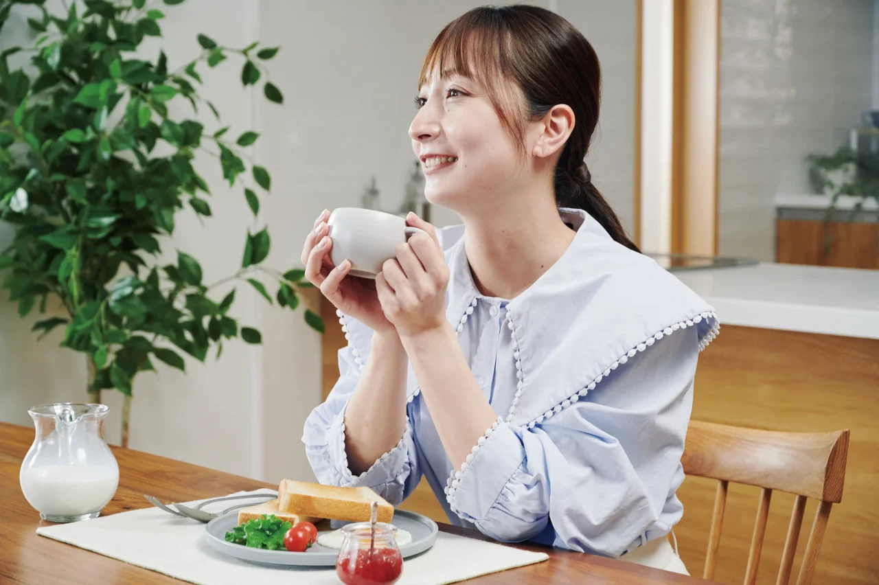白いカップを両手で持ち微笑む女性。テーブルにトーストとミルクピッチャー