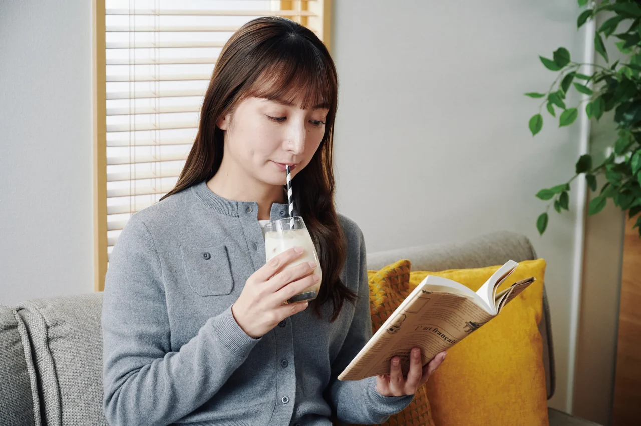 ストローを挿したグラスの白い飲み物を飲みながら本を読む女性