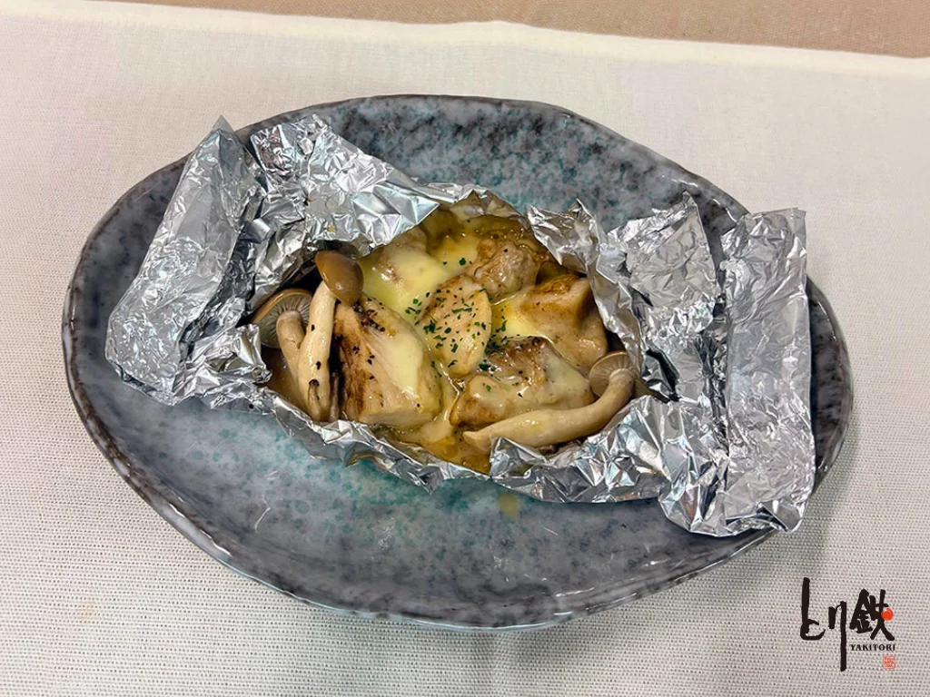 奥州美鶏ときのこのホイルチーズ焼き。皿の上のホイルにチーズと鶏、きのこ