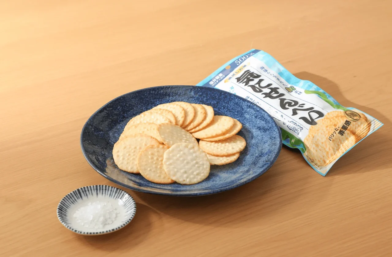 「気にせんべい 満足しお味」の袋と皿に盛ったせんべい、塩の小皿